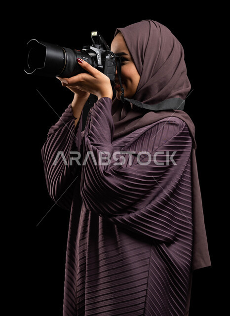 كاميرا عالية الدقة، التقاط الصور التذكارية، بورتريه مقرب من الجانب لإمرأة عربية خليجية سعودية محجبة ترتدي العباءة وتقوم بالتقاط الصور الفوتوغرافية، توثيق اللحظات وتخليدها في الذاكرة، استكشاف العالم من خلال العدسة، تقنيات التصوير المبتكرة، خلفية سوداء.