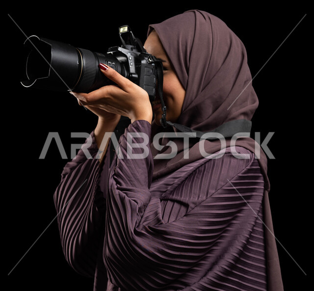 التقاط الصور التذكارية، بورتريه مقرب من الجانب لإمرأة عربية خليجية سعودية محجبة ترتدي العباءة وتقوم بالتقاط الصور الفوتوغرافية، توثيق اللحظات وتخليدها في الذاكرة، كاميرا عالية الدقة، استكشاف العالم من خلال العدسة، تقنيات التصوير المبتكرة، خلفية سوداء.