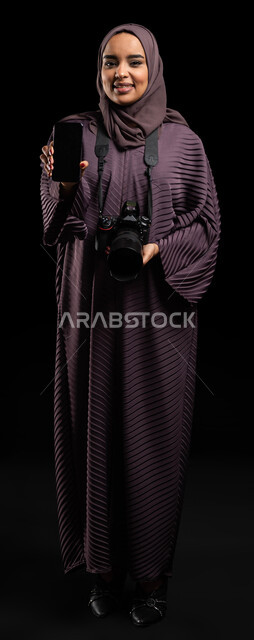 استخدام تطبيقات الهاتف المحمول لتعديل الصور الفوتوغرافية، تقنيات التصوير المبتكرة، بورتريه لإمرأة عربية خليجية سعودية محجبة ترتدي العباءة وتحمل بيدها آلة التصوير والهاتف المحمول بشاشة سوداء فارغه، صورة الجسم بالطول الكامل، خلفية سوداء.