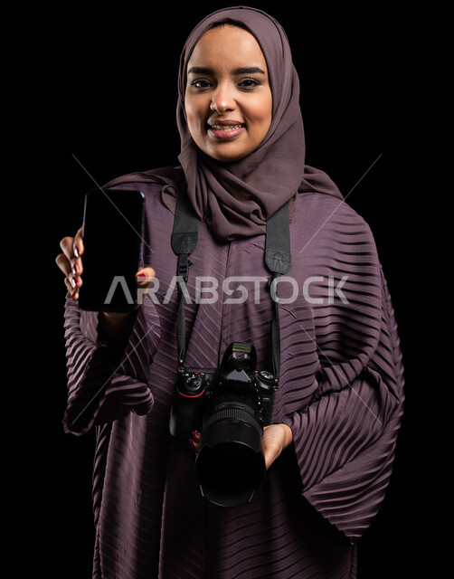 تعديل الصور الفوتوغرافية من خلال تطبيقات الهاتف المحمول، بورتريه مقرب لإمرأة عربية خليجية سعودية محجبة ترتدي العباءة وتحمل بيدها آلة التصوير والهاتف المحمول بشاشة سوداء فارغه، توثيق اللحظات وتخليدها في الذاكرة، تقنيات التصوير المبتكرة، خلفية سوداء.