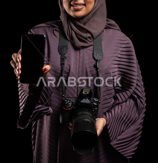 Innovative photography techniques, editing photographs through mobile phone applications, a close-up portrait of a veiled Saudi Arabian Gulf woman wearing an abaya and holding a camera and mobile phone in her hand with a blank black screen, documenting moments and immortalizing them in memory, black background.