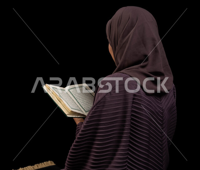 التدبر والخشوع في العبادة، التقرب الى الله تعالى، بورتريه من الخلف لإمرأه عربيه خليجية سعودية محجبة ترتدي العباءة تجلس على سجادة الصلاة وتقرأ القرآن الكريم، المناسبات الدينية والأعياد الإسلامية، التعبد في بيوت الله،  ايماءات الهدوء والاندماج، خلفية سوداء.