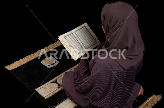 التقرب الى الله تعالى، بورتريه من الخلف لإمرأه عربيه خليجية سعودية محجبة ترتدي العباءة تجلس على سجادة الصلاة وتقرأ القرآن الكريم، المناسبات الدينية والأعياد الإسلامية، التعبد في بيوت الله، التدبر والخشوع في العبادة، ايماءات الهدوء والاندماج، خلفية سوداء.