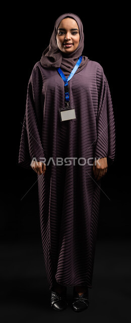 ايماءات الثقة بالنفس، بطاقة شخصية للطلاب والموظقين, التعبير عن الذات، إبراز الهوية الشخصية، بورتريه لإمرأه عربيه خليجية سعودية محجبة مبتسمه ترتدي العباءة وتضع بطاقة بيضاء فارغه بشريط أزرق وتنظر الى الكاميرا، صورة الجسم بالطول الكامل، خلفية سوداء
