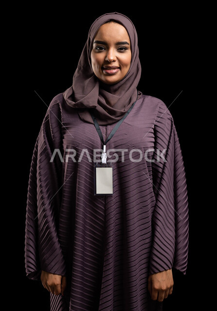 بطاقة شخصية للطلاب والموظقين, التعبير عن الذات، إبراز الهوية الشخصية، بورتريه مقرب لإمرأه عربيه خليجية سعودية محجبة مبتسمه ترتدي العباءة وتضع بطاقة بيضاء فارغه بشريط أسود وتنظر الى الكاميرا، ايماءات الثقة بالنفس، خلفية سوداء