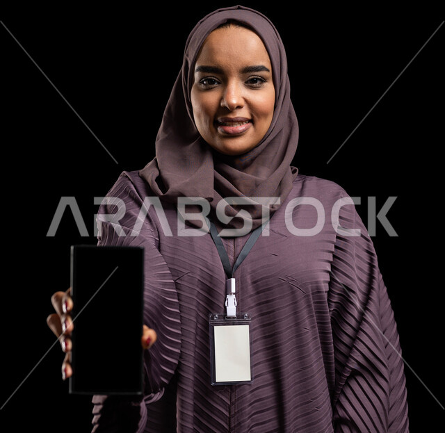 ايماءت الاعجاب باستخدام التقنيه والتكنولوجيا الحديثه،  التسويق الإلكتروني وعرض  وتطبيقات الجوال، إستخدام برامج الهاتف الذكي، بورتريه مقرب لإمرأه عربيه خليجية سعودية محجبة مبتسمه ترتدي العباءة وتضع بطاقة بيضاء بشريط أسود تمسك الهاتف المحمول بشاشة سوداء فارغة، خلفية سوداء