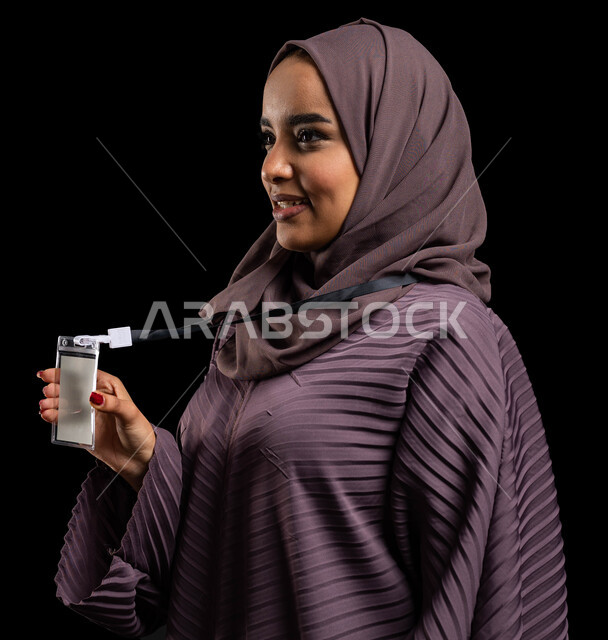 الهوية الشخصية والتعبير عن الذات، بادج شخصي لطالبة جامعية, بورتريه مقرب لإمرأه عربيه خليجية سعودية محجبة مبتسمه ترتدي العباءة وتمسك بيدها بطاقتها الشخصية، بطاقة بيضاء فارغه بشريط أسود، ايماءات الثقة بالنفس، خلفية سوداء