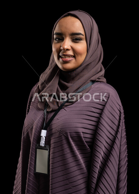 بادج شخصي لطالبة جامعية, بورتريه مقرب لإمرأه عربيه خليجية سعودية محجبة مبتسمه تضع بطاقة بيضاء فارغه بشريط ازرق، بطاقة تعريفية للطلاب والموظفين, ايماءات الثقة بالنفس, الهوية الشخصية والتعبير عن الذات، خلفية سوداء