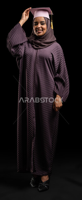 Celebrating the completion of a university degree, the transition from academic life to the labor market, education in the Kingdom of Saudi Arabia, portrait of a university student, a Saudi Arabian Gulf graduate wearing an abaya and holding a graduation cap in her hand, full-length body portrait, black background