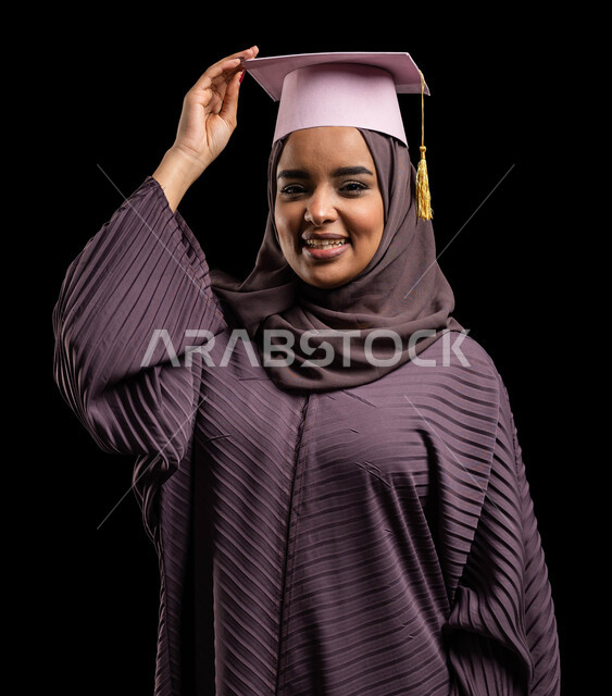 الاحتفالات بتحقيق حلم النجاح من الجامعة، لحظة تاريخية في حياة الخريجين، جودة التعليم في جامعات المملكة العربية السعودية, بورتريه مقرب لطالبة جامعية خريجة عربية سعودية خليجية ترتدي العباءة وتمسك بيدها قبعة التخرج ، خلفية سوداء