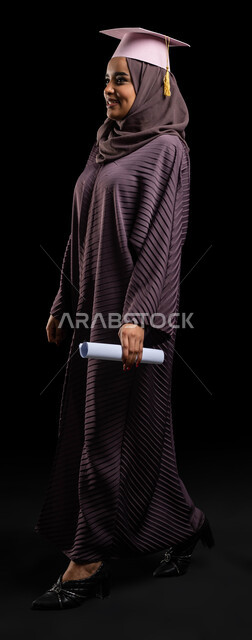 التخرج وتحقيق الذات، التعليم في المملكة العربية السعودية,  بورتريه لطالبة جامعية خريجة عربية سعودية خليجية ترتدي العباءة وقبعة التخرج وتمسك بيدها شهادة التخرج، السرور والفرح والاحتفال بيوم التخرج، صورة الجسم بالطول الكامل، خلفية سوداء