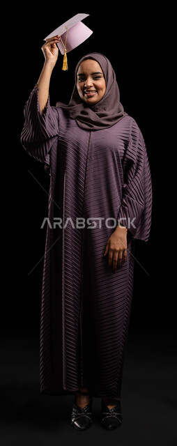 التخرج والتحضير للحياة المهنية، الانتقال من الحياة الأكاديمية إلى سوق العمل، بورتريه  لطالبة جامعية خريجة عربية سعودية خليجية ترتدي العباءة وتمسك بيدها قبعة التخرج، الاحتفالات بتحقيق حلم النجاح من الجامعة، صورة الجسم بالطول الكامل، خلفية سوداء
