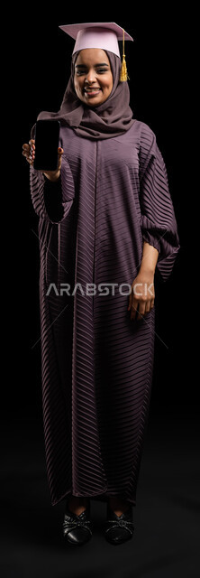 التقديم على وظائف ومهن في السعودية، التجهيز لحفلات التخرج عبر التطبيقات الالكترونية، بورتريه لطالبة جامعية خريجة عربية سعودية خليجية ترتدي العباءة وقبعة التخرج وتمسك بيدها الهاتف المحمول، عرض شاشه سوداء فارغة، صورة الجسم بالطول الكامل، خلفية سوداء.