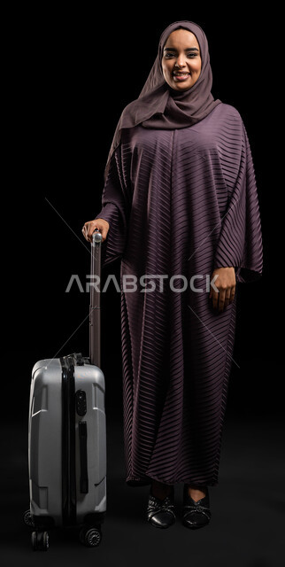 متعة الاستعداد والتجهيز للسفر، بورتريه لإمرأة عربية خليجية سعودية محجبة ترتدي العباءة تمسك حقيبة السفر، مفهوم السياحة والفندقة، ايماءات النشاط والحيوية ، قضاء أوقات ممتعة خارج البلاد، صورة الجسم بالطول الكامل، خلفية سوداء.