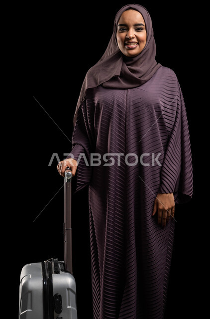 قضاء أوقات مسلية خارج البلاد، متعة الاستعداد للرحلة السياحية، بورتريه لإمرأة عربية خليجية سعودية محجبة ترتدي العباءة تمسك حقيبة السفر، مفهوم السياحة والفندقة، ايماءات الحماس لرحلة ممتعه، خلفية سوداء.