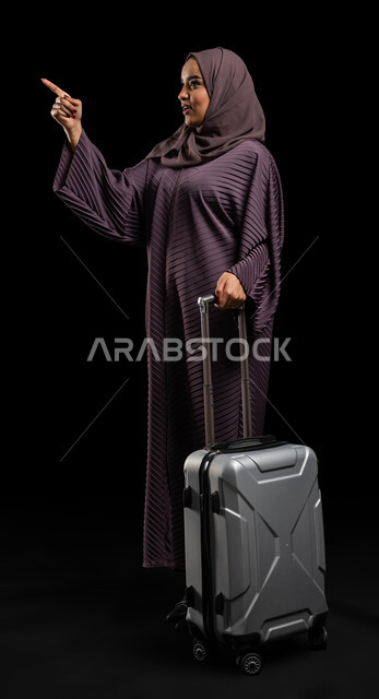 قضاء أوقات ممتعة ومسلية في الإجازة، بورتريه لشابة خليجية سعودية محجبة ترتدي العباءة الخليجية تمسك بيدها حقيبة السفر، تشير بإصبع السبابة إلى شيء ما، الاستعداد لرحلة سياحية، صورة الجسم بالطول الكامل، خلفية سوداء،