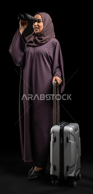 رؤية الأشياء البعيدة عبر المنظار، حب الاستكشاف والمغامرة، بورتريه لإمرأة عربية خليجية سعودية محجبة ترتدي العباءة الخليجية تحمل بيدها منظار ثنائي العينين( التلسكوب) وتمسك حقيبة السفر، قضاء أوقات ممتعة في الإجازة، الاستعداد للرحلة السياحية، صورة الجسم بالطول الكامل، خلفية سوداء.