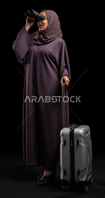 Seeing distant things through binoculars, love of exploration and adventure, portrait of a veiled Saudi Arabian Gulf woman wearing the Gulf abaya, holding a pair of binoculars (telescope) in her hand and holding a travel bag, spending enjoyable times on vacation, preparing for a tourist trip, full-length body portrait, black background.