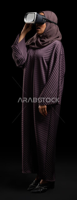The concept of modern electronic technologies, gestures of interaction and integration with holographic elements, a close-up portrait of a veiled Saudi Gulf Arab woman sitting on a chair wearing an abaya and 3D virtual reality (VR) glasses, full-length body image, black background.