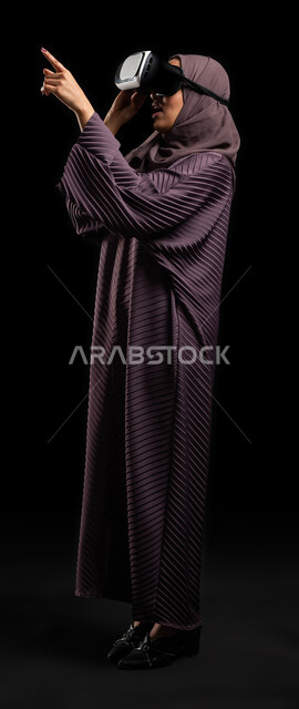 التفاعل والاندماج باستخدام حاسة اللمس لتحريك العناصر الهولوجرامية، بورتريه لامرأة عربية خليجية سعودية محجبة تجلس على كرسي ترتدي العباءة ونظارات الواقع الإفتراضي (في آر) ثلاثية الأبعاد تشير بإصبعها السبابة لشيء ما، مفهوم التقنيات الإلكترونية الحديثة، صورة الجسم بالطول الكامل، خلفية سوداء.