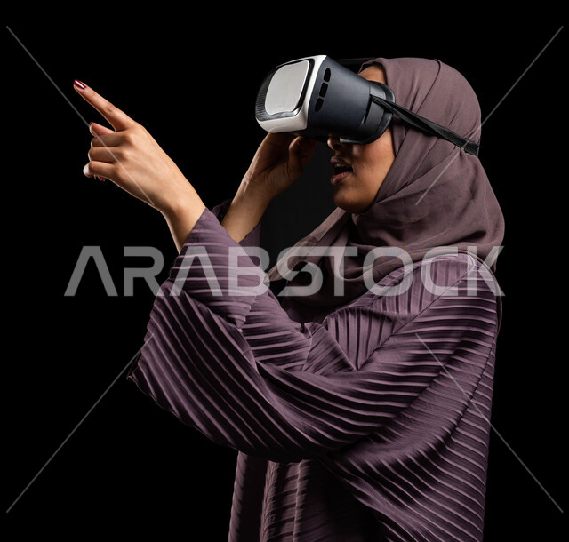 The concept of modern electronic technologies, interaction and integration using the sense of touch to move holographic elements, a close-up portrait of a veiled Saudi Gulf Arab woman sitting on a chair wearing an abaya and 3D virtual reality (VR) glasses, pointing with her index finger at something, black background.