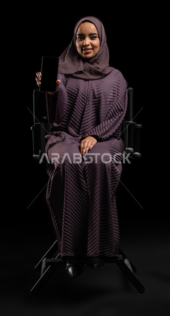 Displaying mobile phone applications, electronic marketing using technology and technology, a portrait of a smiling Saudi Gulf Arab woman wearing the hijab and abaya, holding a mobile phone with a blank black screen in her hand, looking at the camera, black background.