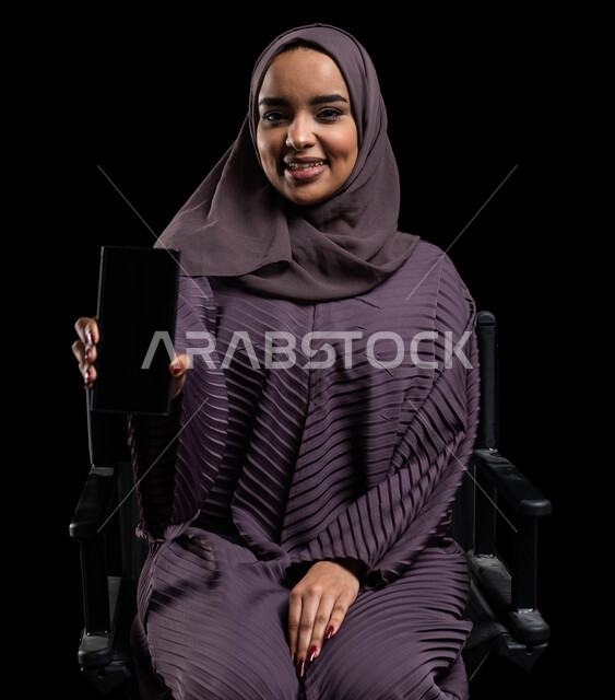 التسويق الالكتروني باستخدام التقنية والتكنولوجيا، عرض تطبيقات الهاتف المحمول، بورتريه مقرب لشابة عربية خليجية سعودية مبتسمة ترتدي الحجاب والعباءة تحمل بيدها جوال بشاشة سوداء فارغة، النظر الى الكاميرا، خلفية سوداء.