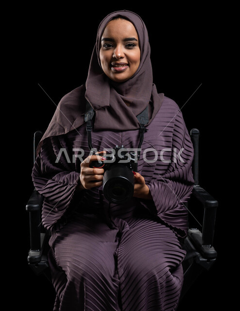 Facial gestures indicating happiness, taking souvenir photos using a photography camera, talent and professionalism in the art of photography with a high-resolution camera, a portrait of a veiled Saudi Gulf Arab young woman wearing an abaya, holding a camera in her hand, black background.