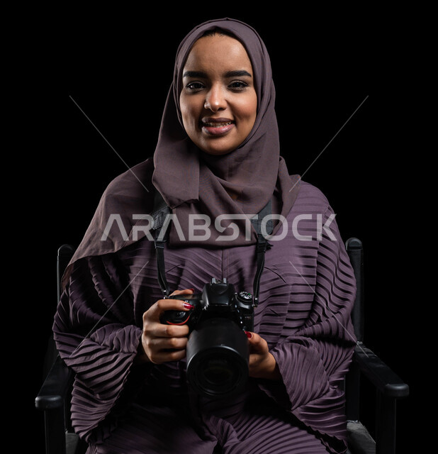 إيماءات وجه تدل على السرور، التقاط الصور التذكارية باستخدام كاميرا التصوير الفوتوغرافي، موهبة واحتراف فن التصوير بكاميرا عالية الدقة، بورتريه مقرب لشابة عربية خليجية سعودية محجبة ترتدي عباءة تحمل بيدها كاميرا، خلفية سوداء.