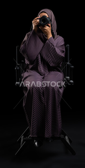 Talent and professionalism in the art of photography with a high-resolution camera, a portrait of a veiled Saudi Arabian Gulf woman wearing an abaya and holding a camera in her hand, taking souvenir photos using a photography camera, black background.