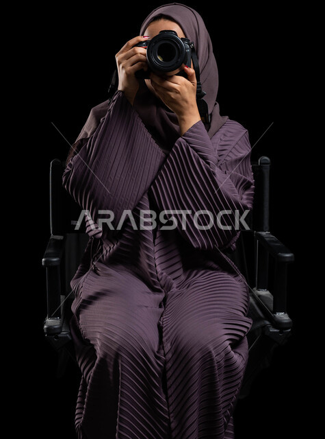 موهبة واحتراف فن التصوير بكاميرا عالية الدقة، بورتريه لشابة عربية خليجية سعودية محجبة ترتدي عباءة تحمل بيدها كاميرا، تلتقط الصور التذكارية باستخدام كاميرا التصوير الفوتوغرافي، خلفية سوداء.