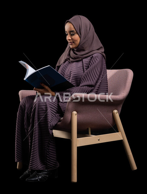 إنجاز الواجبات اليومية, قضاء الوقت بالقراءة والتعلم والتثقف ، بورتريه لامرأة عربية سعودية خليجية محجبة ترتدي العباءة تجلس على كرسي مريح وتحمل بيديها كتاب، الاستمتاع بقراءة الكتب والقصص والروايات, ثقافة التعلم الذاتي, خلفية سوداء.