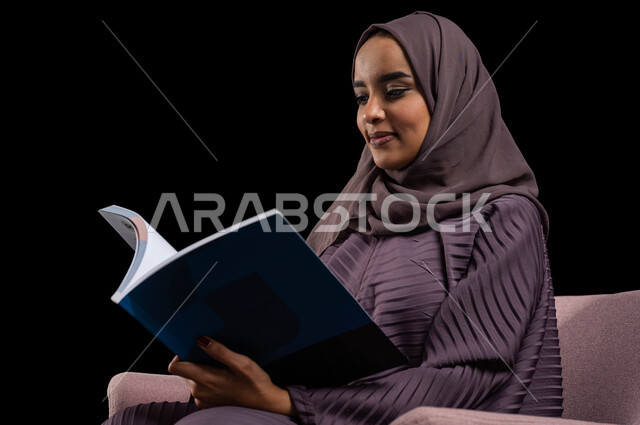Enjoying reading books, stories and novels, completing daily duties, spending time reading, learning and becoming acculturated, a close-up portrait of a veiled Saudi Arabian Gulf woman wearing an abaya, sitting on a comfortable chair and holding a book in her hands, the culture of self-learning, black background.