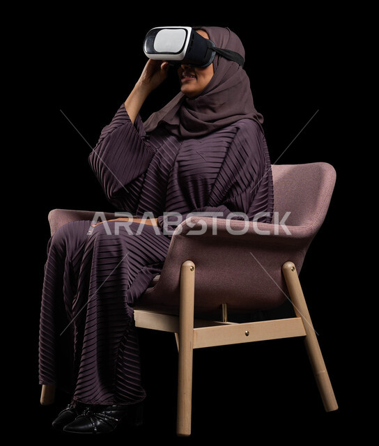 وسائل ترفيهية في وقت الفراغ, الاستمتاع بمشاهدة أفلام الواقع الافتراضي، بورتريه لإمرأة عربية خليجية سعودية تجلس على كرسي مريح وترتدي نظارة الواقع الافتراضي ثلاثية الأبعاد، قضاء أوقات ممتعة ومسلية بالألعاب الافتراضية، خلفية سوداء.