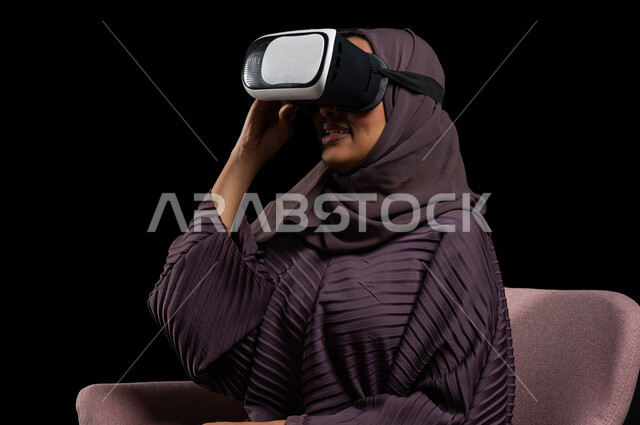 قضاء أوقات ممتعة ومسلية بالألعاب الافتراضية، وسائل ترفيهية في وقت الفراغ, الاستمتاع بمشاهدة أفلام الواقع الافتراضي، بورتريه مقرب لإمرأة عربية خليجية سعودية تجلس على كرسي مريح وترتدي نظارة الواقع الافتراضي ثلاثية الأبعاد، خلفية سوداء.