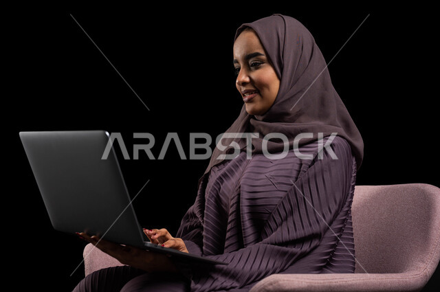 Working remotely and browsing the Internet using modern technologies, shopping on websites, a close-up portrait of a veiled Saudi Arabian Gulf woman wearing an abaya, sitting on a chair using a laptop computer, chatting, socializing, and making a video call, black background.