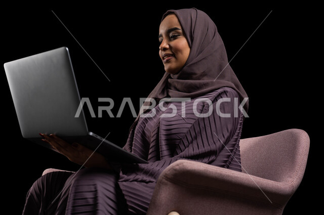 العمل عن بعد وتصفح الانترنت باستخدام تقنيات حديثة، التسوق عبر المواقع الإلكترونية، بورتريه مقرب لشابة عربية خلجية سعودية محجبة ترتدي عباءة تجلس على كرسي تستخدام الحاسوب المحمول, الدردشة والتواصل الاجتماعي وإجراء مكالمة فيديو, خلفية سوداء.