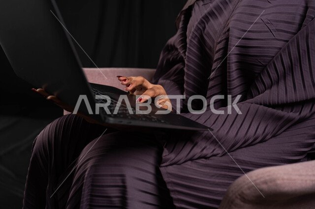 Working remotely and browsing the Internet using modern technologies, shopping on websites, a close-up portrait of a veiled Saudi Arabian Gulf woman wearing an abaya, sitting on a chair using a laptop computer, chatting, socializing, and making a video call, black background.