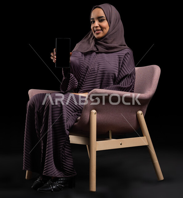 بث مباشر ودردشة ومحادثات مع الأقارب عن بعد، بورتريه لإمرأة عربية خليجية سعودية محجبة ترتدي العباءة تجلس على كرسي تقوم بالتواصل مع الاصدقاء عبر الهاتف المحمول، ايماءات الاندماج والسرور بإجراء مكالمات الفيديو، مفهوم الجلوس والراحة، خلفية سوداء.