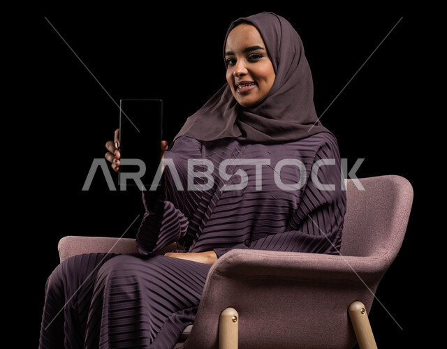 بث مباشر ودردشة ومحادثات مع الأقارب عن بعد، بورتريه لإمرأة عربية خليجية سعودية محجبة ترتدي العباءة تجلس على كرسي تقوم بالتواصل مع الاصدقاء عبر الهاتف المحمول، ايماءات الاندماج والسرور بإجراء مكالمات الفيديو، مفهوم الجلوس والراحة، خلفية سوداء.