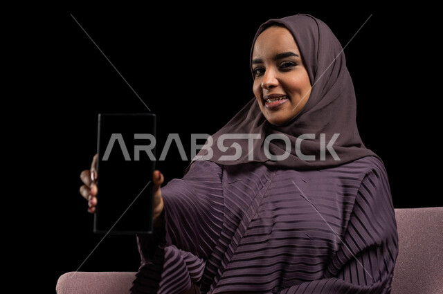 دردشة ومحادثات مع الأقارب عن بعد، بورتريه مقرب لإمرأة عربية خليجية سعودية محجبة ترتدي العباءة تجلس على كرسي تقوم بالتواصل مع الاصدقاء عبر الهاتف المحمول، ايماءات الاندماج والسرور بإجراء مكالمات الفيديو، مفهوم الجلوس والراحة، خلفية سوداء.