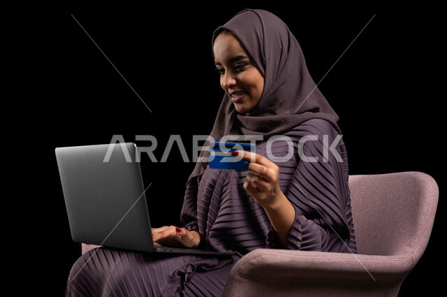 إمكانية الشراء والدفع من خلال البرامج والتطبيقات الالكترونية، استخدام أجهزة تقنية حديثة للدفع عن بعد عبر الانترنت، بورتريه مقرب لسيدة عربية خليجية سعودية محجبة ترتدي عباءة تجلس على كرسي وتحمل الحاسوب المحمول وبطاقة الإئتمان البنكية للتسوق الالكتروني، خلفية سوداء.