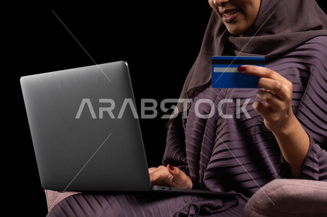 إمكانية الشراء والدفع من خلال البرامج والتطبيقات الالكترونية، استخدام أجهزة تقنية حديثة للدفع عن بعد عبر الانترنت، بورتريه مقرب لسيدة عربية خليجية سعودية محجبة ترتدي عباءة تجلس على كرسي وتحمل الحاسوب المحمول وبطاقة الإئتمان البنكية للتسوق الالكتروني، خلفية سوداء.