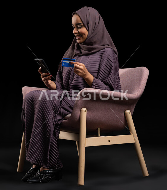 إمكانية الشراء والدفع من خلال برامج وتطبيقات عبر الجوال، بورتريه لسيدة عربية خليجية سعودية محجبة ترتدي عباءة تجلس على كرسي وتحمل بيدها الهاتف المحمول وبطاقة الإئتمان البنكية للتسوق الالكتروني، استخدام أجهزة تقنية حديثة، الدفع عن بعد عبر الانترنت، خلفية سوداء.
