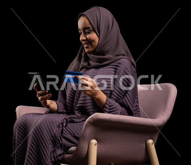 إمكانية الشراء والدفع من خلال برامج وتطبيقات عبر الجوال، بورتريه لسيدة عربية خليجية سعودية محجبة ترتدي عباءة تجلس على كرسي وتحمل بيدها الهاتف المحمول وبطاقة الإئتمان البنكية للتسوق الالكتروني، استخدام أجهزة تقنية حديثة، الدفع عن بعد عبر الانترنت، خلفية سوداء.