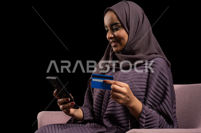 استخدام أجهزة تقنية حديثة للدفع عن بعد عبر الانترنت، إمكانية الشراء والدفع من خلال برامج وتطبيقات عبر الجوال، بورتريه مقرب لسيدة عربية خليجية سعودية محجبة ترتدي عباءة تجلس على كرسي وتحمل بيدها الهاتف المحمول وبطاقة الإئتمان البنكية للتسوق الالكتروني، خلفية سوداء.