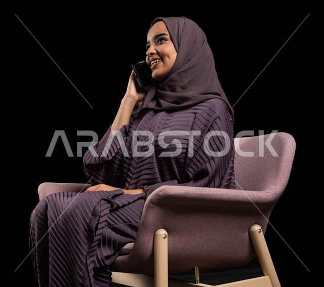 التواصل مع الأقارب عن بعد، بورتريه لإمرأة عربية خليجية سعودية محجبة ترتدي العباءة تجلس على كرسي تقوم بالتواصل مع الاصدقاء عبر الهاتف المحمول، ايماءات الاندماج والسرور بإجراء المكالمات الهاتفية، مفهوم الجلوس والراحة، خلفية سوداء.