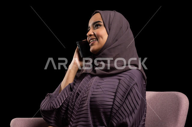 مفهوم الجلوس والراحة، التواصل مع الأقارب عن بعد، بورتريه مقرب لإمرأة عربية خليجية سعودية محجبة ترتدي العباءة تجلس على كرسي تقوم بالتواصل مع الاصدقاء عبر الهاتف المحمول، ايماءات الاندماج والسرور بإجراء المكالمات الهاتفية، خلفية سوداء.