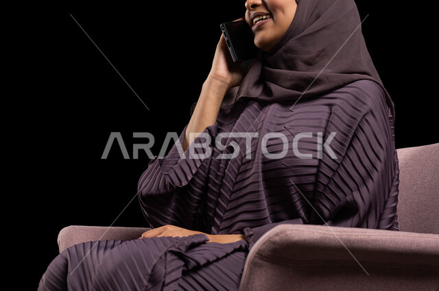 مفهوم الجلوس والراحة، التواصل مع الأقارب عن بعد، بورتريه مقرب لإمرأة عربية خليجية سعودية محجبة ترتدي العباءة تجلس على كرسي تقوم بالتواصل مع الاصدقاء عبر الهاتف المحمول، ايماءات الاندماج والسرور بإجراء المكالمات الهاتفية، خلفية سوداء.