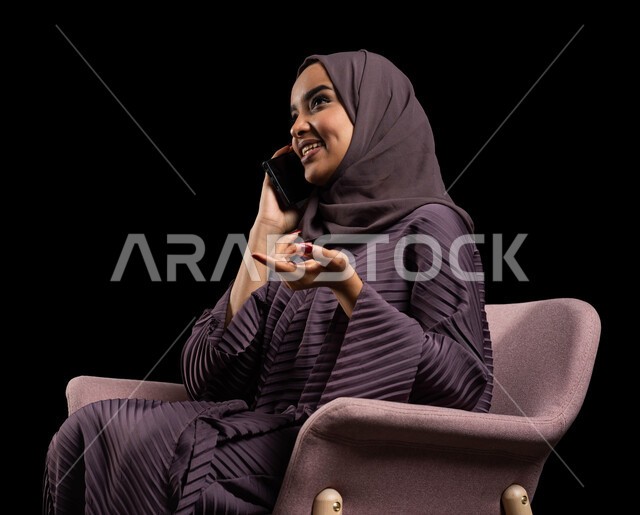 مفهوم الجلوس والراحة، التواصل مع الأقارب عن بعد، بورتريه مقرب لإمرأة عربية خليجية سعودية محجبة ترتدي العباءة تجلس على كرسي تقوم بالتواصل مع الاصدقاء عبر الهاتف المحمول، ايماءات الاندماج والسرور بإجراء المكالمات الهاتفية، خلفية سوداء.